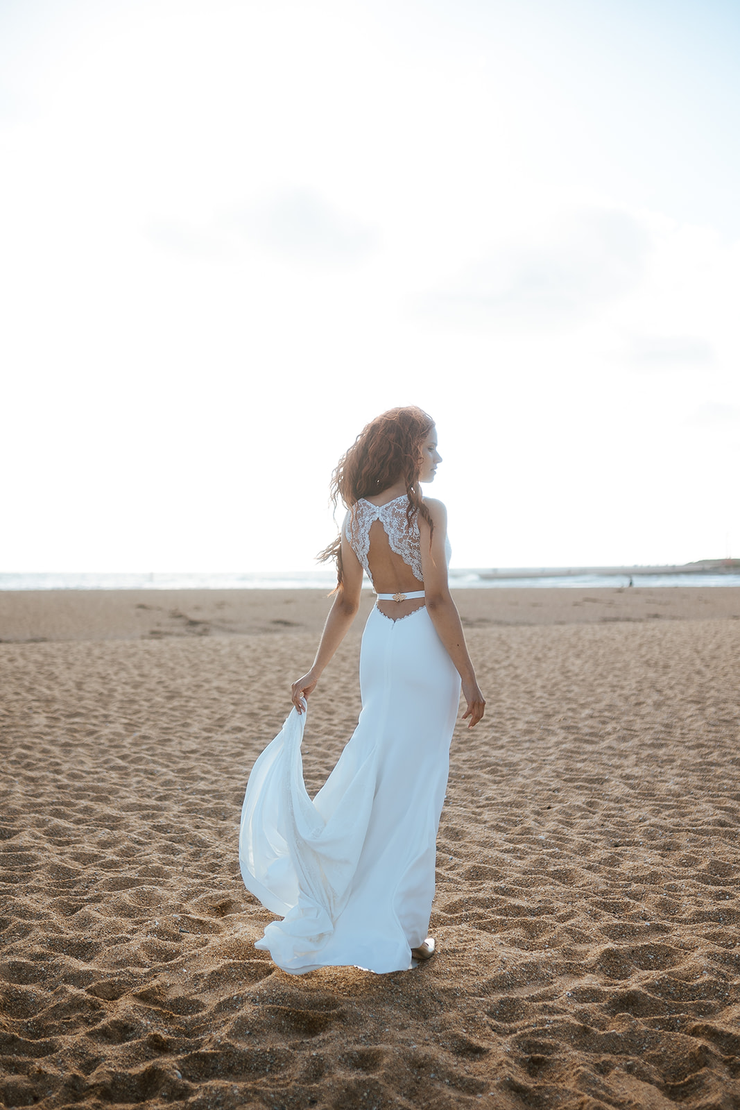 Femme rousse portant une robe de mariée de créateur en dentelle sur-mesure à la plage.